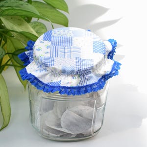 May include: A glass jar filled with tea bags, topped with a blue and white patchwork fabric lid with a blue lace trim.