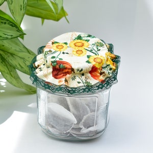 Peut inclure: Un bocal en verre avec un couvercle en tissu floral rempli de sachets de thé. Le couvercle est orné d'une dentelle verte et présente un motif floral avec des tournesols et des citrouilles.