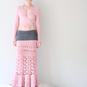 May include: A pink crochet skirt with a gray waistband and a matching pink top. The skirt features an openwork design and a flared hem. The top is a long-sleeved wrap style in a light pink color. The outfit is a bohemian style.