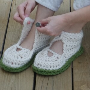 Op de afbeelding: Handgemaakte gehaakte pantoffels in gebroken wit met een groene zool. De pantoffels hebben een Mary Jane-stijl met een bandje en een knoopsluiting. Het dikke garen zorgt voor een textuur, perfect voor binnenshuis.