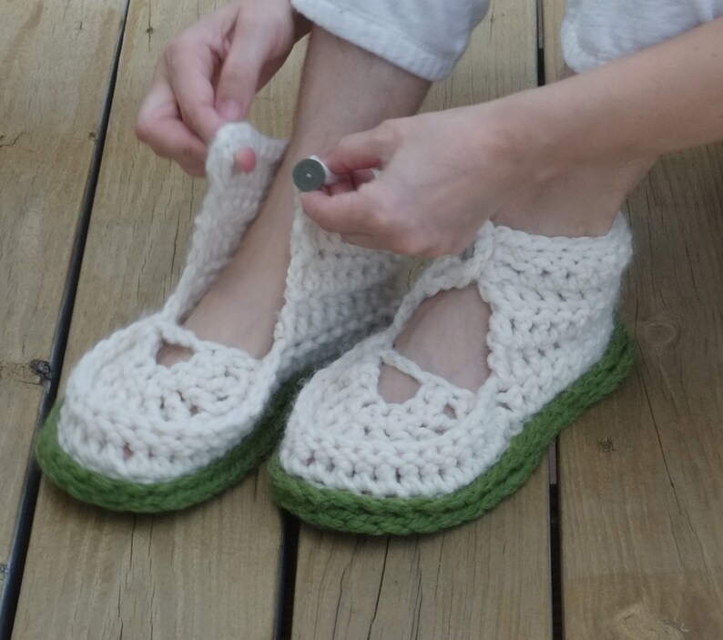 Op de afbeelding: Handgemaakte gehaakte pantoffels in gebroken wit met een groene zool. De pantoffels hebben een Mary Jane-stijl met een bandje en een knoopsluiting. Het dikke garen zorgt voor een textuur, perfect voor binnenshuis.