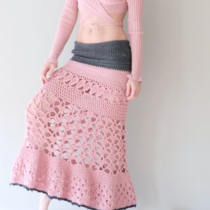 May include: A handmade, floor-length crochet skirt in shades of pink and gray. The skirt features a wide gray waistband, transitioning to a pink, open-weave design. The bottom hem is trimmed with a dark gray border. The skirt is a bohemian style.