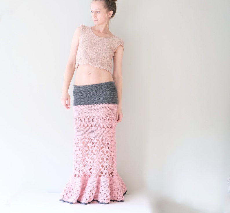 May include: A handmade, crochet skirt in a mermaid style. The skirt features a gray waistband, a pink body with openwork detailing, and a flared hem. The skirt is paired with a light pink lace crop top. The overall look is bohemian and feminine.