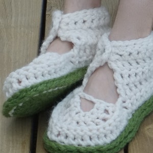 Op de afbeelding: Een paar handgemaakte, cr&egrave;mekleurige gehaakte pantoffels met een groene zool en rand. De pantoffels hebben een Mary Jane-stijl met een bandje over de bovenkant van de voet en een uitgesneden detail. Het dikke garen geeft ze een gezellige, getextureerde uitstraling.