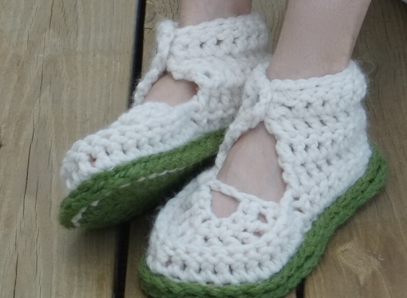 Op de afbeelding: Een paar handgemaakte, cr&egrave;mekleurige gehaakte pantoffels met een groene zool en rand. De pantoffels hebben een Mary Jane-stijl met een bandje over de bovenkant van de voet en een uitgesneden detail. Het dikke garen geeft ze een gezellige, getextureerde uitstraling.