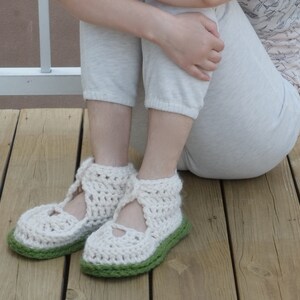 Op de afbeelding: Handgemaakte gehaakte pantoffels in een cr&egrave;mekleur met een groene zool. De pantoffels hebben een Mary Jane-stijl met een bandje over de bovenkant van de voet. Het open weefsel zorgt voor ademend vermogen en comfort. Deze gezellige pantoffels zijn perfect voor binnenshuis.