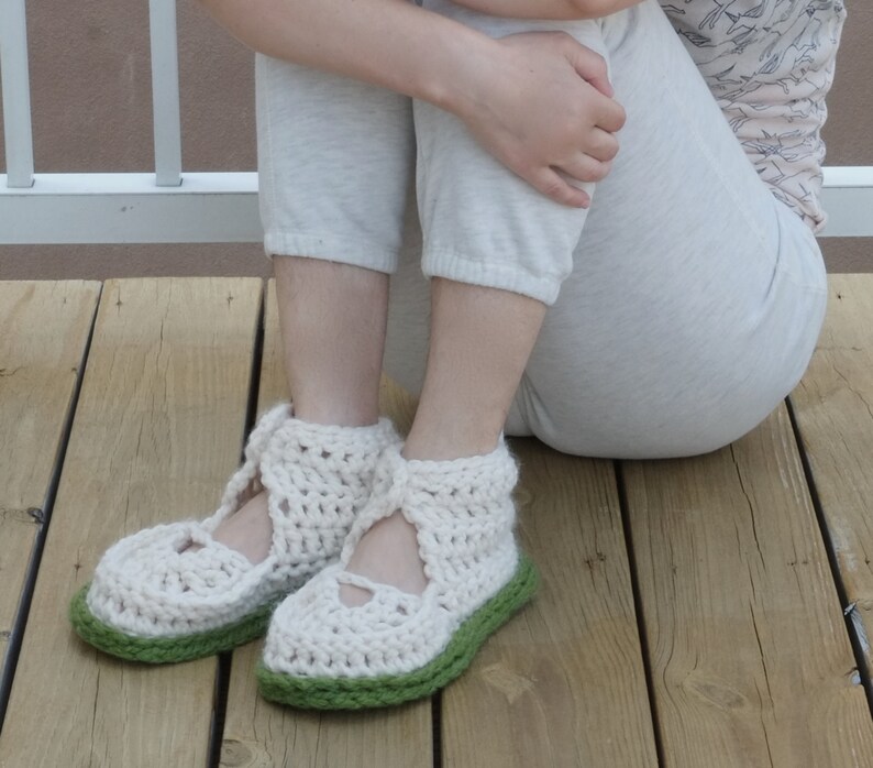 Op de afbeelding: Handgemaakte gehaakte pantoffels in een cr&egrave;mekleur met een groene zool. De pantoffels hebben een Mary Jane-stijl met een bandje over de bovenkant van de voet. Het open weefsel zorgt voor ademend vermogen en comfort. Deze gezellige pantoffels zijn perfect voor binnenshuis.