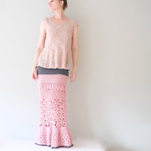 May include: A light pink lace top paired with a long, handmade crochet skirt. The skirt features a gray waistband, a pink patterned midsection, and a flared bottom. The crochet work creates an open, airy design.