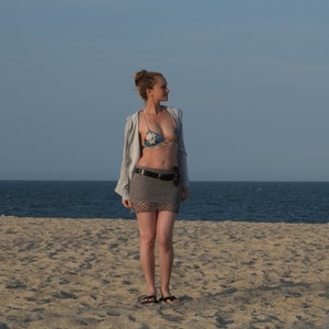 May include: A woman wearing a floral bikini top, gray crochet skirt, and a gray jacket stands on a sandy beach. She is also wearing a black belt and sandals. The ocean and sky are visible in the background.