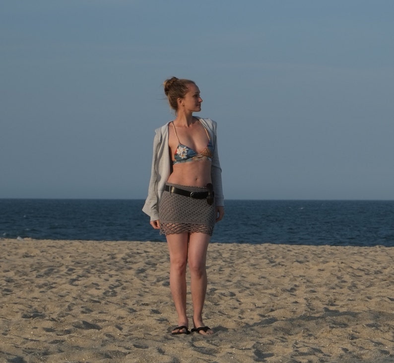 May include: A woman wearing a floral bikini top, gray crochet skirt, and a gray jacket stands on a sandy beach. She is also wearing a black belt and sandals. The ocean and sky are visible in the background.
