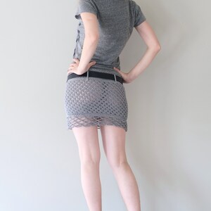 May include: A gray crochet mini skirt with a scalloped hem, worn with a black belt. The skirt is made of a loosely woven material, revealing the legs. The person is wearing a gray t-shirt.