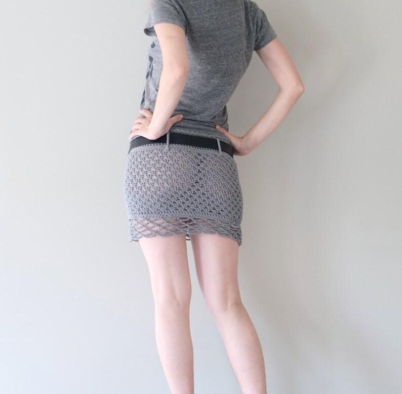 May include: A gray crochet mini skirt with a scalloped hem, worn with a black belt. The skirt is made of a loosely woven material, revealing the legs. The person is wearing a gray t-shirt.
