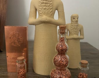 Könnte beinhalten: Drei kleine Glasflaschen gefüllt mit Kupferflocken, neben zwei kleinen beigefarbenen Statuen und einem Kupferbarren. Der Kupferbarren trägt die Aufschrift "COPPER 999 FINE".