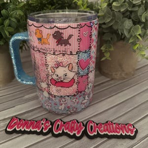 May include: A tumbler with a blue glitter handle and a clear lid. The tumbler features a patchwork design with images of cats, hearts, and the text "Everybody wants to be a cat." The bottom of the tumbler has a blue and white pattern.