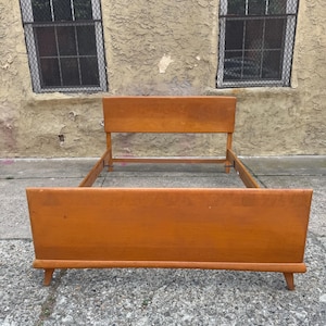 May include: A mid-century modern bed frame constructed from light brown wood. The piece features a rectangular headboard and a matching footboard, both with clean, straight lines. The frame is supported by four tapered wooden legs.