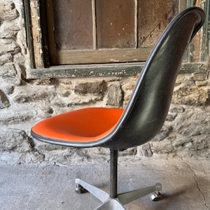 May include: An orange and black office chair with a five-point base and wheels. The seat is orange, and the back is black. The chair has a metal base and a curved back. The chair is set against a stone wall.