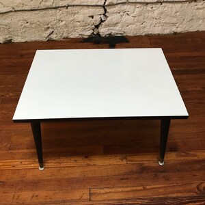 May include: A white square coffee table with black legs. The table is sitting on a wooden floor.