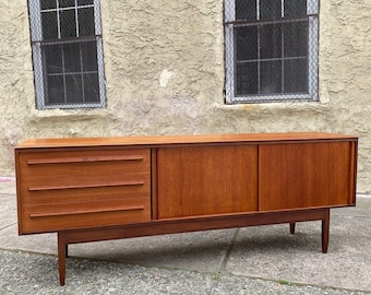 May include: A mid-century modern teak sideboard with three drawers and two doors. The sideboard has a warm, honey-colored finish and sits on four tapered legs.