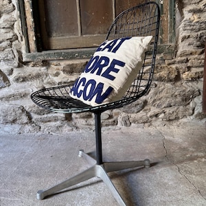May include: A black wire chair with a cushion that reads "EAT MORE BACON" in navy blue letters. The chair has a black stem and a silver, four-legged base. The background features a stone wall and a window.