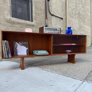 May include: Mid-century modern wooden cabinet with a warm brown finish. The cabinet features open shelving and a lower section supported by angled legs. Items on display include a vintage radio, a blue glass vase, and a small typewriter.