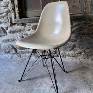 May include: A beige molded plastic chair with a curved seat and back. The chair has a black metal wire base with four legs. The chair is set against a stone wall and concrete floor.