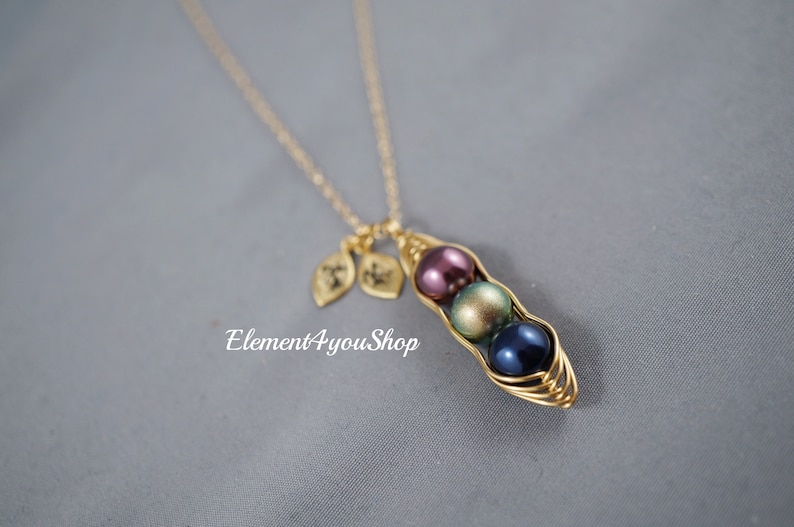 May include: A gold-colored necklace featuring a pea pod pendant with three colorful pearl beads: purple, green, and blue. Two leaf charms hang near the clasp. The text "Element4youShop" is visible.
