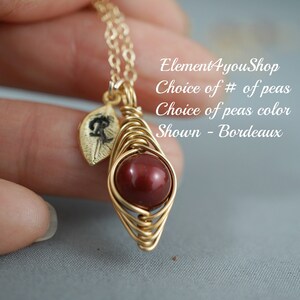 May include: A gold-colored wire-wrapped pendant necklace with a round, deep red bead. A small gold leaf charm with a black design hangs next to the bead. The text on the image says "Choice of # of peas" and "Shown - Bordeaux".