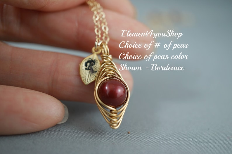 May include: A gold-colored wire-wrapped pendant necklace with a round, deep red bead. A small gold leaf charm with a black design hangs next to the bead. The text on the image says "Choice of # of peas" and "Shown - Bordeaux".
