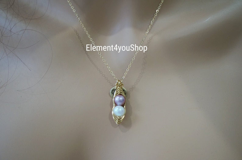 May include: A gold-colored necklace with a pendant shaped like a pea pod. The pea pod contains two pearls, one lavender and one white. The necklace chain is delicate. The text "Element4youShop" is visible.