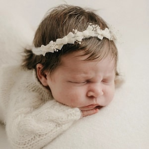 Può includere: Un neonato che indossa un maglione di lana bianco e una corona di fiori bianchi con piccoli fiocchi bianchi.