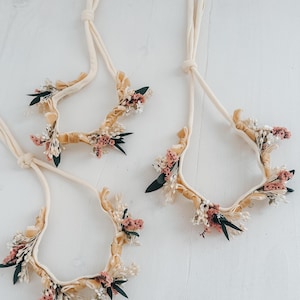 Puede incluir: Cuatro diademas florales con tela color crema y adornadas con flores secas en tonos rosa, blanco y verde. Las diademas est&aacute;n dispuestas sobre una superficie blanca, mostrando su delicado dise&ntilde;o y est&eacute;tica natural.