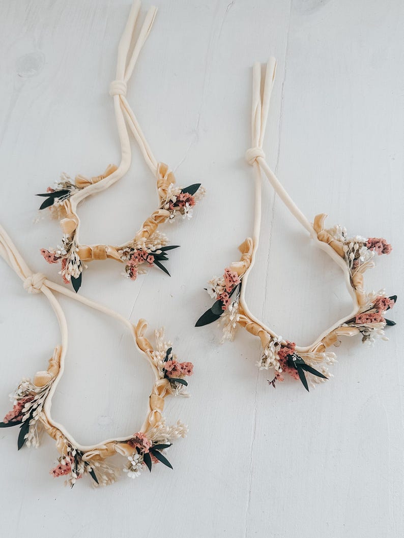 Puede incluir: Cuatro diademas florales con tela color crema y adornadas con flores secas en tonos rosa, blanco y verde. Las diademas est&aacute;n dispuestas sobre una superficie blanca, mostrando su delicado dise&ntilde;o y est&eacute;tica natural.