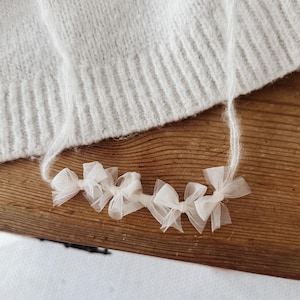 Può includere: Una collana delicata caratterizzata da una serie di piccoli fiocchi in tulle bianco sporco attaccati a un sottile cordino di filato sfocato color crema. I fiocchi sono disposti in una fila orizzontale, creando un aspetto morbido e materico.