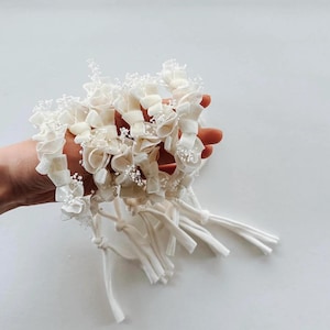 Può includere: Una collezione di accessori per capelli con fiori in tessuto bianco sporco. Ogni pezzo presenta delicati petali a strati e piccoli accenti di perline bianche. Gli accessori sono legati insieme con nastri di tessuto coordinati.