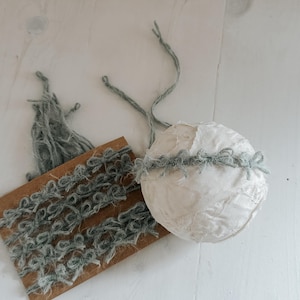 Op de afbeelding: Een witte, getextureerde bol met een grijze garen afwerking en een klein stukje grijs garen bevestigd. Een bruin kartonnen label met grijze garen lussen is ook aanwezig. De items liggen op een wit houten oppervlak.