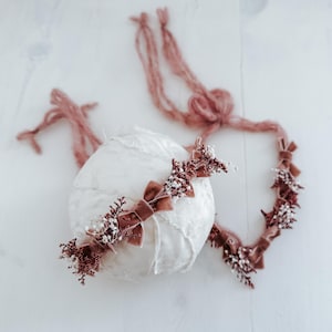 May include: A white, textured sphere is decorated with a dusty rose velvet headband. The headband has small bows and sprigs of dried flowers. Long, matching ties are attached, creating a delicate, bohemian aesthetic.
