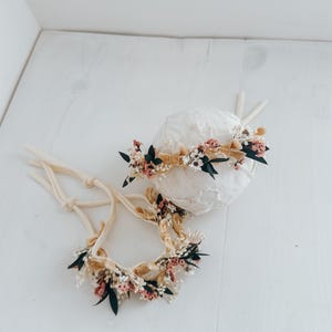 Puede incluir: Dos diademas florales con lazos color crema. Las diademas presentan una variedad de peque&ntilde;as flores en tonos rosa, blanco y verde, acentuadas por una cinta beige. Una diadema est&aacute; sobre una esfera blanca.