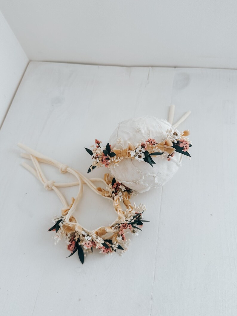 Puede incluir: Dos diademas florales con lazos color crema. Las diademas presentan una variedad de peque&ntilde;as flores en tonos rosa, blanco y verde, acentuadas por una cinta beige. Una diadema est&aacute; sobre una esfera blanca.
