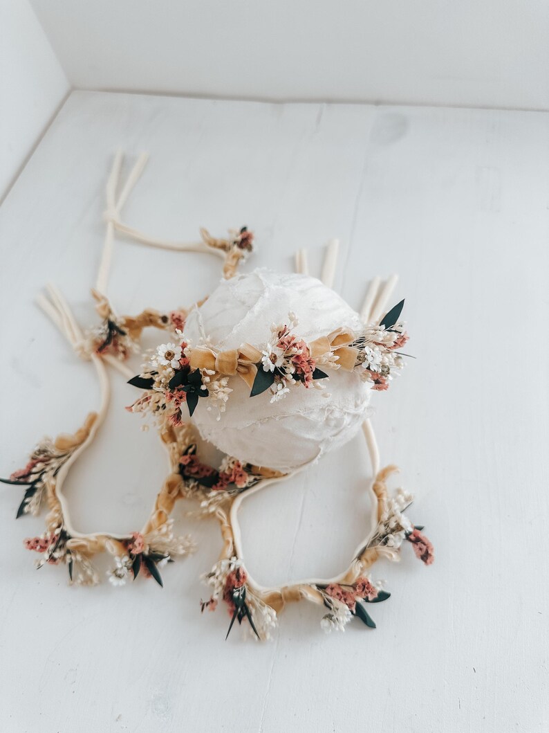 Puede incluir: Un conjunto de diademas florales y una esfera decorativa. Las diademas presentan flores secas en tonos rosa, blanco y verde, acentuadas con cintas beige. La esfera est&aacute; envuelta en tela blanca, creando una est&eacute;tica suave y natural. Ideal para fotograf&iacute;a de reci&eacute;n nacidos.