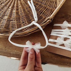 May include: A white elastic headband featuring a small, delicate tulle bow. The headband is made of a soft, stretchy material and is designed to be worn around the head. Additional identical headbands are displayed on a brown cardboard backing.