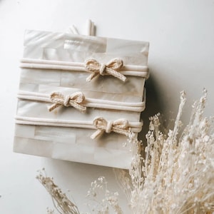 May include: Three white cotton headbands with knotted bows. The headbands are displayed on a white and cream patterned surface.