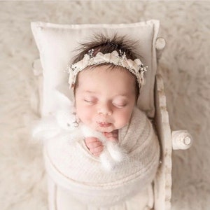 May include: A sleeping newborn baby girl wearing a white headband with a flower design, wrapped in a white blanket, and holding a white stuffed bunny. The baby is lying in a white wooden crib.