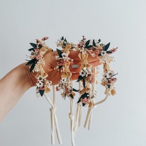 Puede incluir: Una colecci&oacute;n de accesorios florales para el cabello. Cada pieza presenta una base de color crema adornada con peque&ntilde;as flores rosas, blancas y verdes. Los accesorios se mantienen unidos con cordones de color crema, creando una est&eacute;tica delicada y natural.