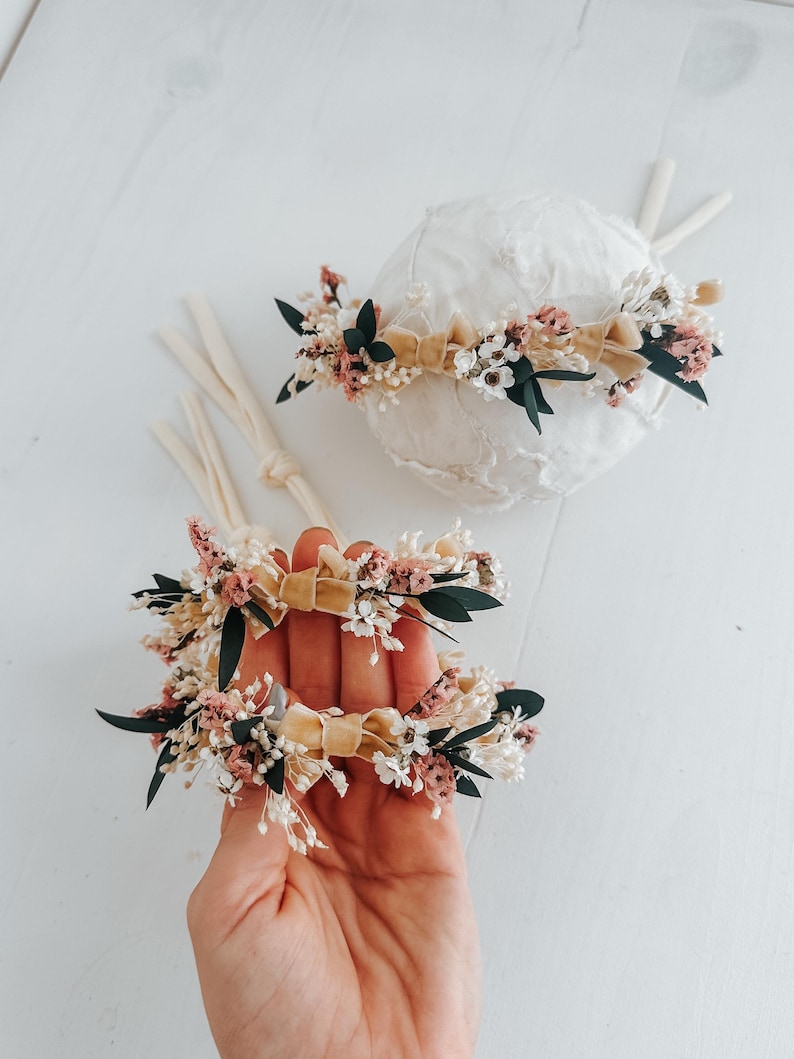 Puede incluir: Dos diademas florales con base de cinta de terciopelo beige. Las diademas est&aacute;n adornadas con peque&ntilde;as flores rosas, blancas y verdes. Una diadema est&aacute; sobre un objeto esf&eacute;rico blanco, y la otra se sostiene en una mano.