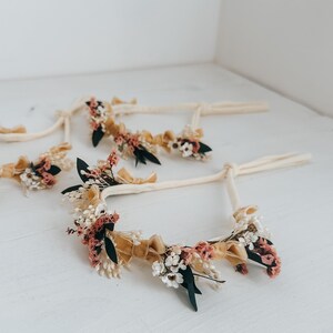 Puede incluir: Varias diademas florales con cintas color crema y adornadas con flores secas en tonos rosa, blanco y verde, acentuadas con cintas doradas. Las diademas est&aacute;n dispuestas sobre una superficie blanca.