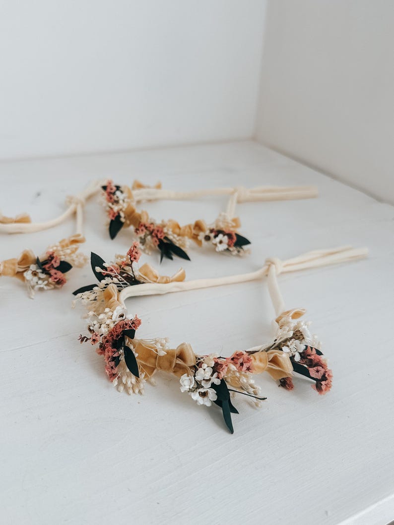 Puede incluir: Varias diademas florales con cintas color crema y adornadas con flores secas en tonos rosa, blanco y verde, acentuadas con cintas doradas. Las diademas est&aacute;n dispuestas sobre una superficie blanca.