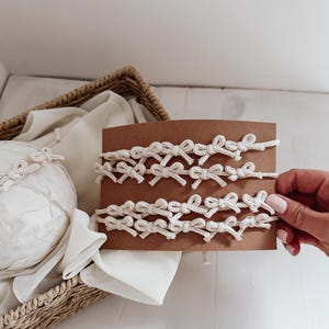 May include: Close-up of several cream-coloured macrame bow bracelets displayed on a brown card. The bracelets are handmade with a series of small bows. A basket and white fabric are in the background.
