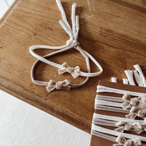 May include: A selection of baby headbands in neutral shades. The headbands have small velvet bows in a light beige colour. Some are tied together, while others are neatly stacked on a brown cardboard display, all on a wooden surface.