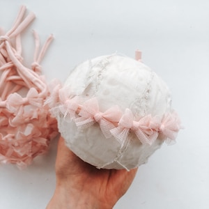 Op de afbeelding: Een witte stoffen bal met een roze tule strik eromheen. De bal is gemaakt van stoffen restjes en heeft een ruwe textuur.