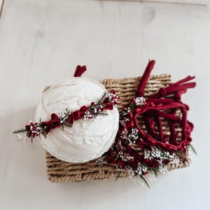 Pode incluir: Uma esfera branca texturizada e duas tiaras vermelhas com detalhes florais repousam sobre uma cesta de vime. As tiaras apresentam veludo vermelho e pequenas flores brancas, sugerindo um tema festivo ou nupcial.